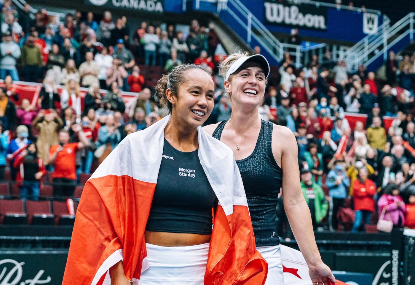 Leylah Fernandez (left) and Gabriela Dabrowski smile on court holding Canadian flags.