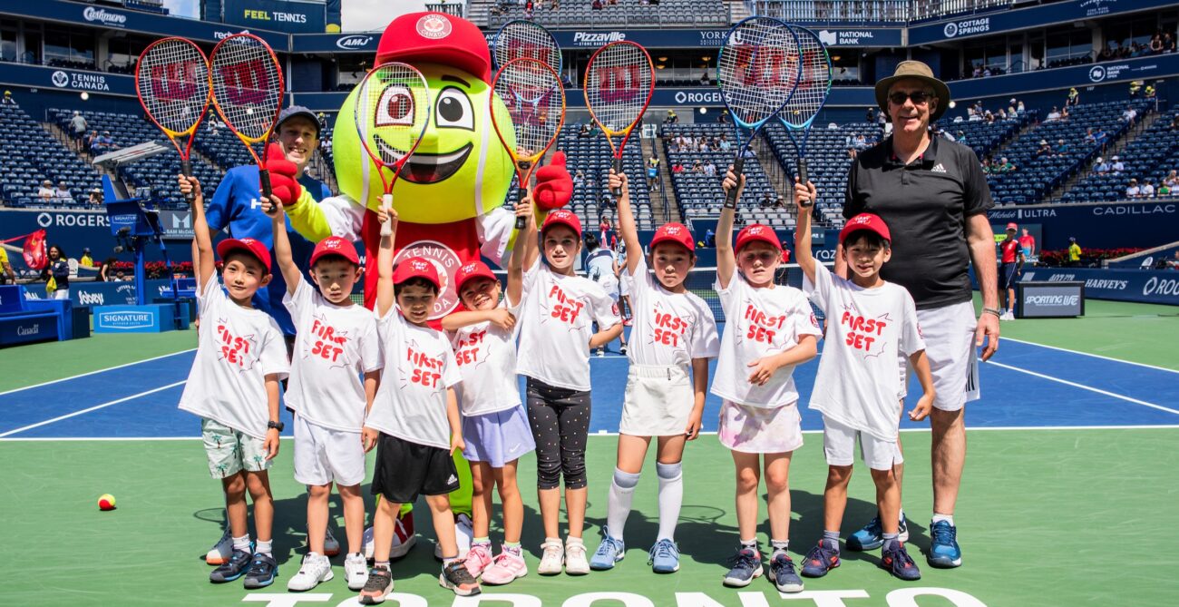 A group of children in the Rogers First Set program stand on Centre Court with the tournament mascot holding racquets in the air.